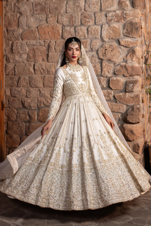 Model wearing full Naqshi Fasana set: dove‑white halter raw‑silk frock, voluminous panels, ornate mirror work, structured pouch & embellished dupatta for bridal elegance.
