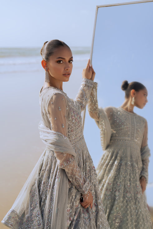 Model wearing full NAQSHI Valeria 3PC: expertly embellished silver net frock with hand‑adda detailing, plain PK raw‑silk pants & matching embroidered dupatta for a refined formal look.