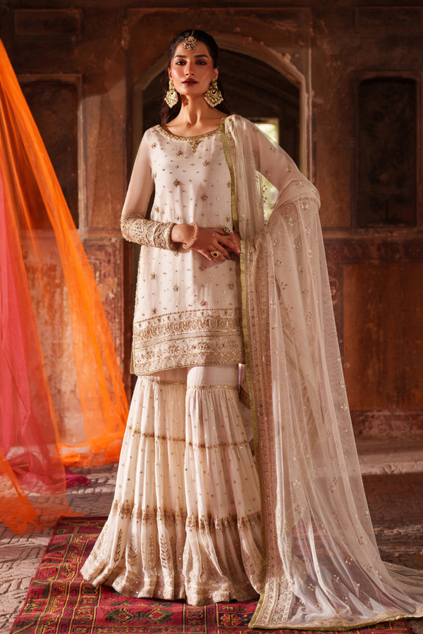 Woman in traditional cream-colored outfit with orange dupatta against a textured wall background