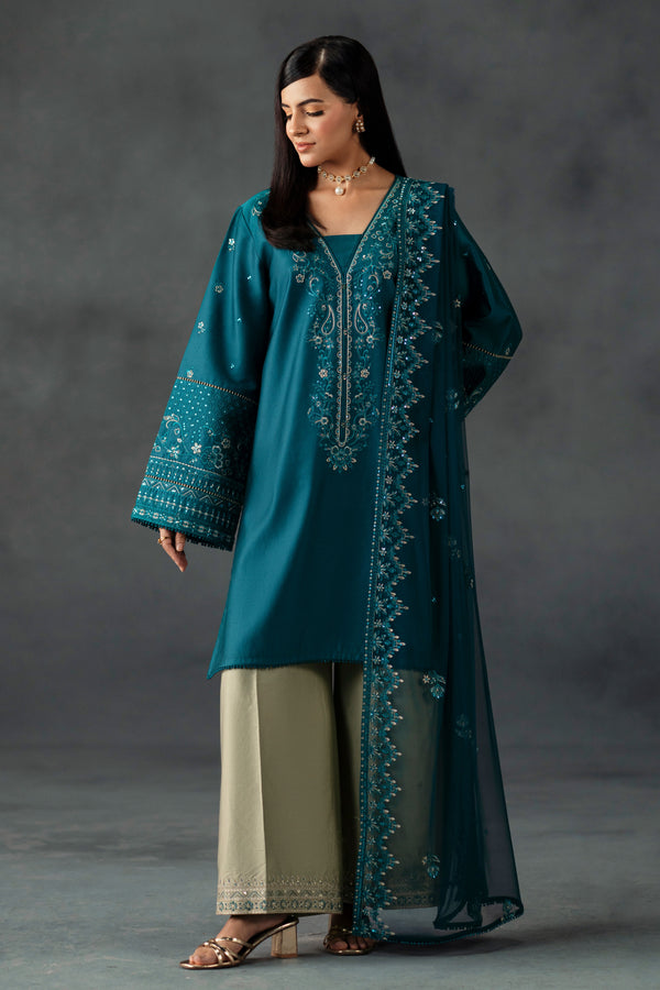 Woman wearing a teal embroidered outfit against a gray background