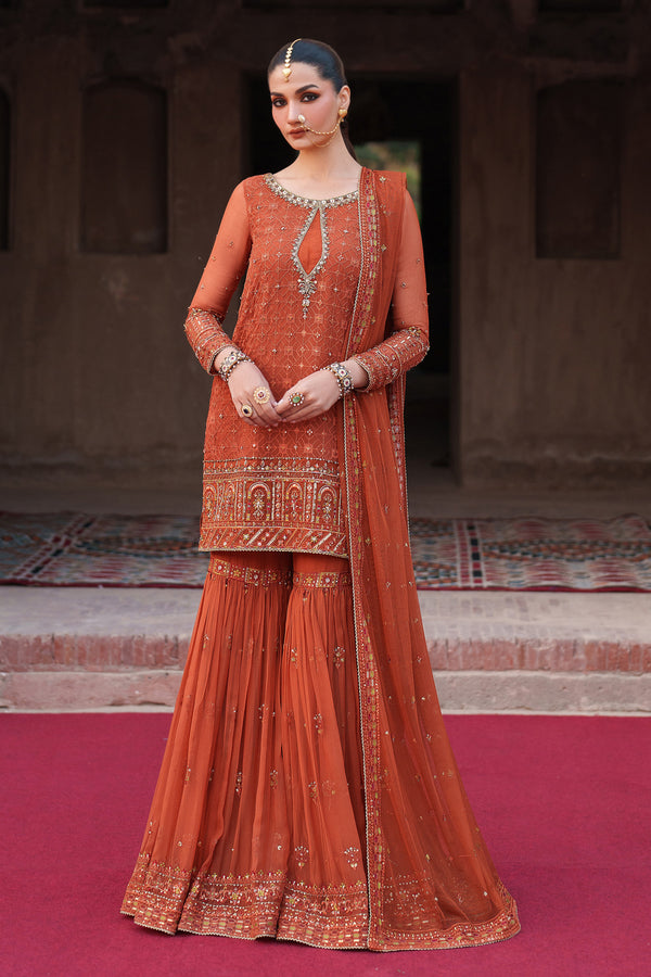 Woman in an orange traditional outfit with intricate patterns standing on a red carpet.