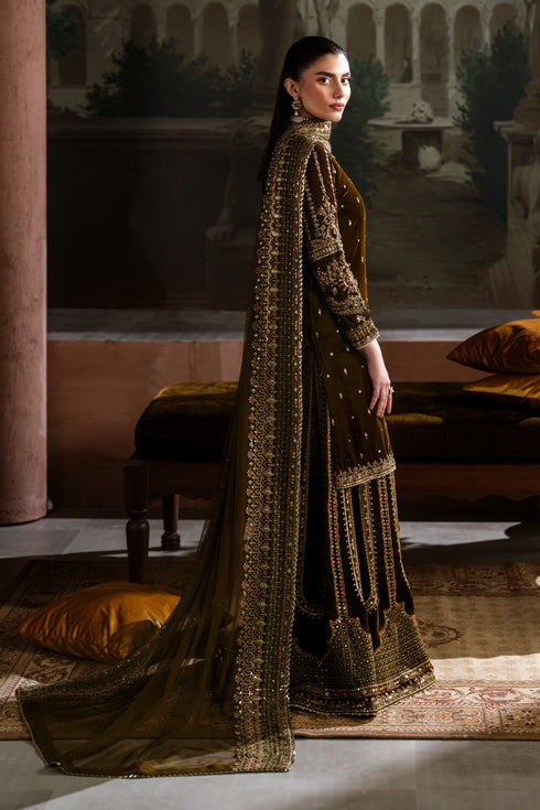 Woman in a long, dark green embroidered dress standing in an indoor setting with classical architecture.