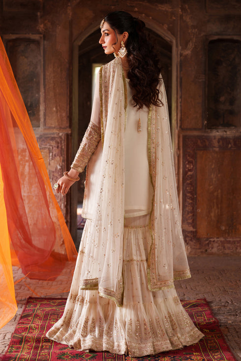 Woman in traditional white and gold outfit with orange dupatta in a decorative setting