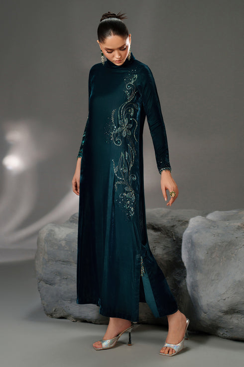 Woman wearing a long, dark green dress with intricate patterns standing against a gray background.