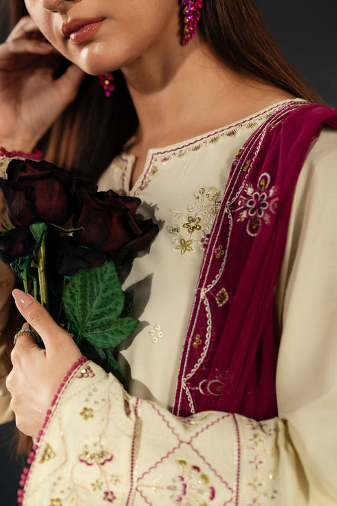 Woman wearing a traditional embroidered outfit with a purple shawl, holding dark flowers.