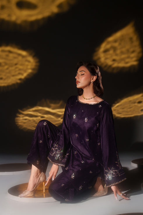 Woman in a dark purple traditional outfit with gold embroidery sitting on a white surface with a dark background.