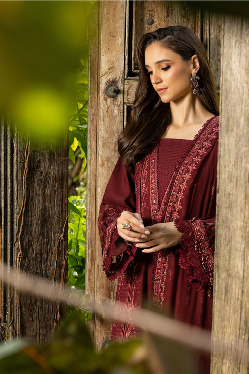 Woman in a burgundy embroidered dress standing in a wooden doorway with greenery around.