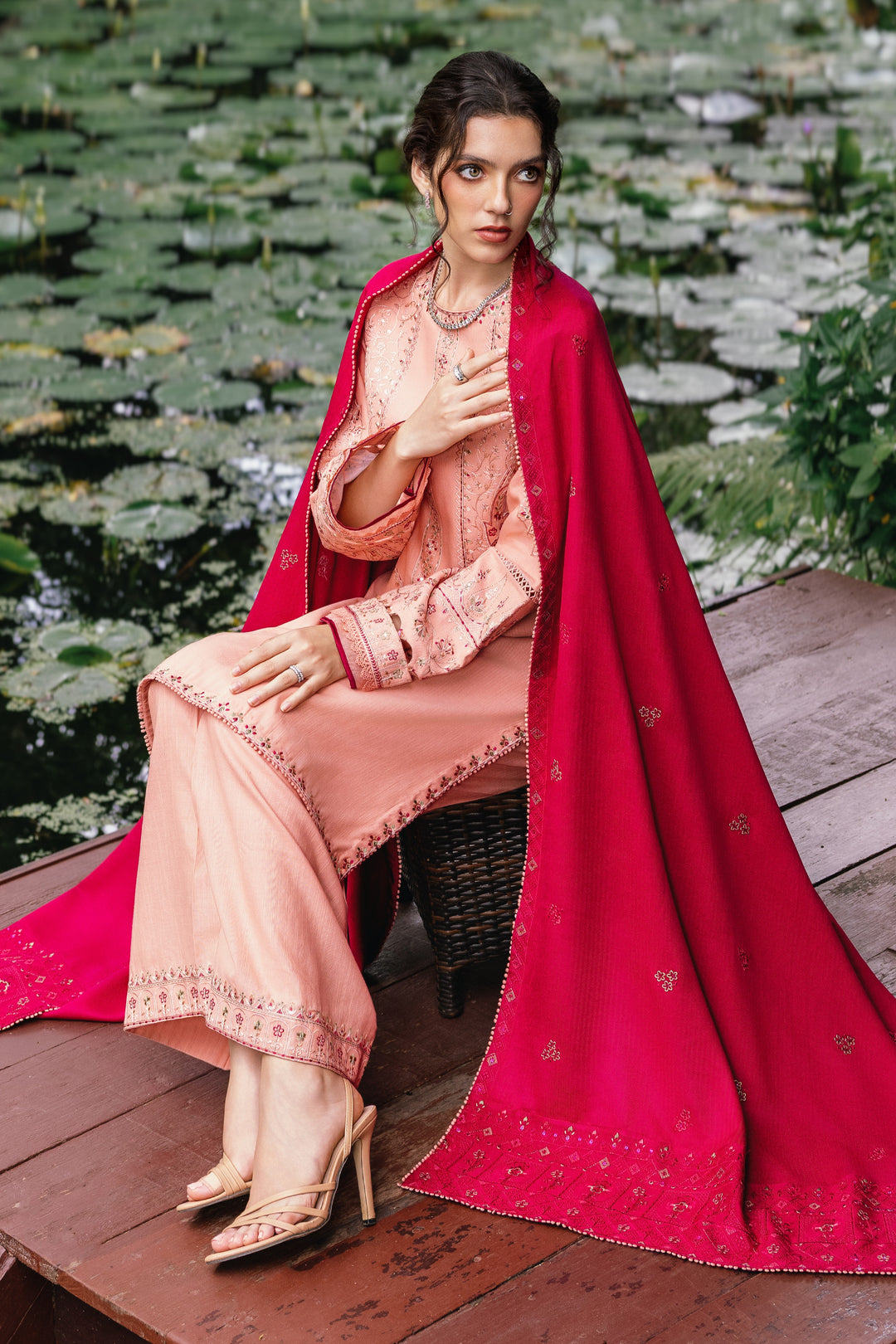 Woman in a peach outfit with a red dupatta sitting on a wooden platform with water lilies in the background.