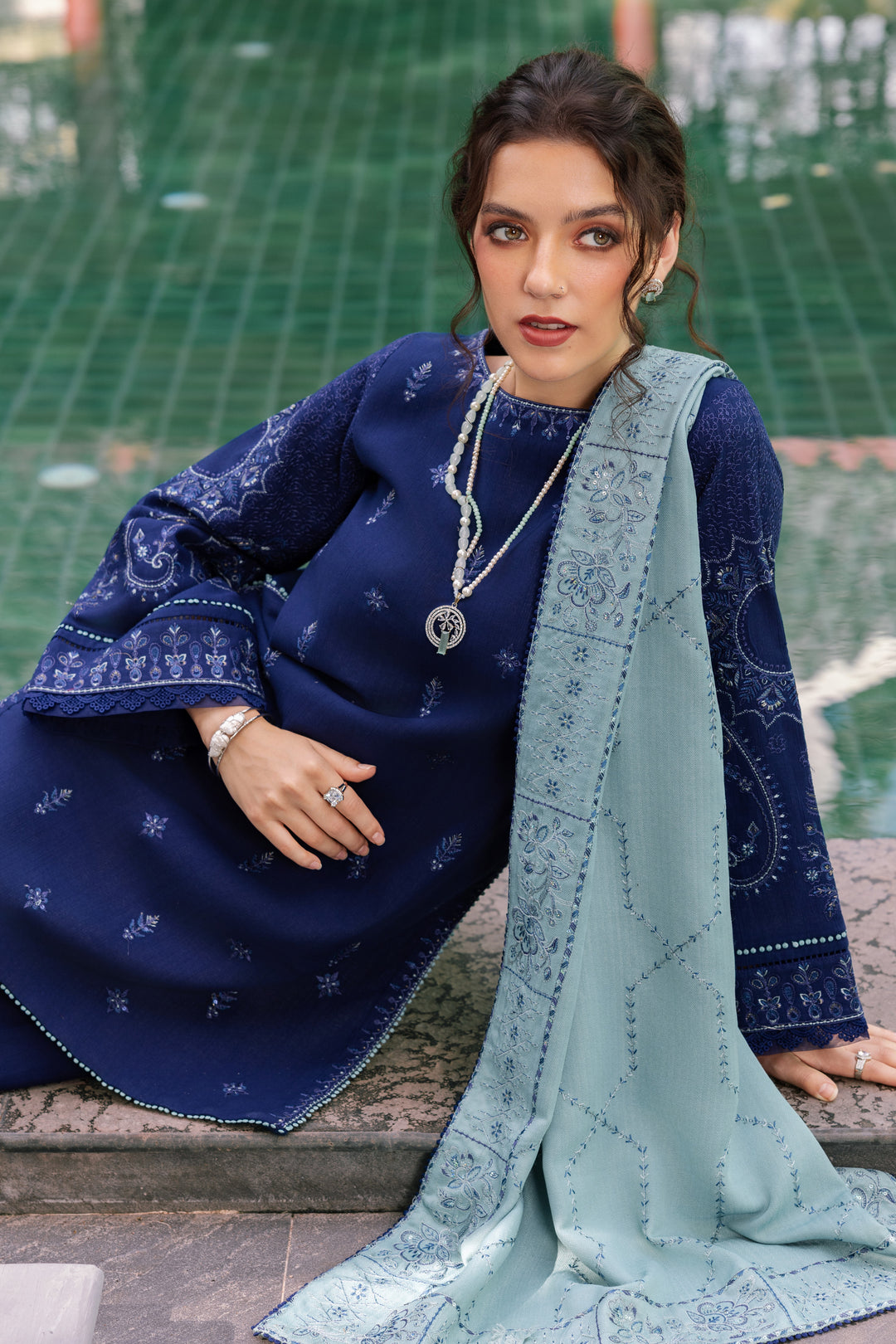 Woman in traditional blue and green outfit with jewelry, sitting on a stone bench.