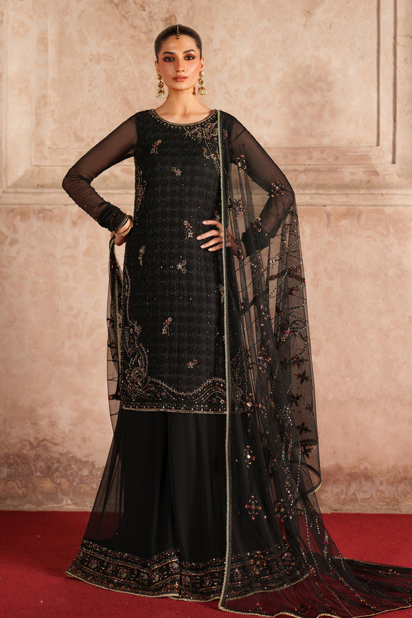 Woman in a black embroidered traditional outfit with a sheer dupatta against a beige wall.