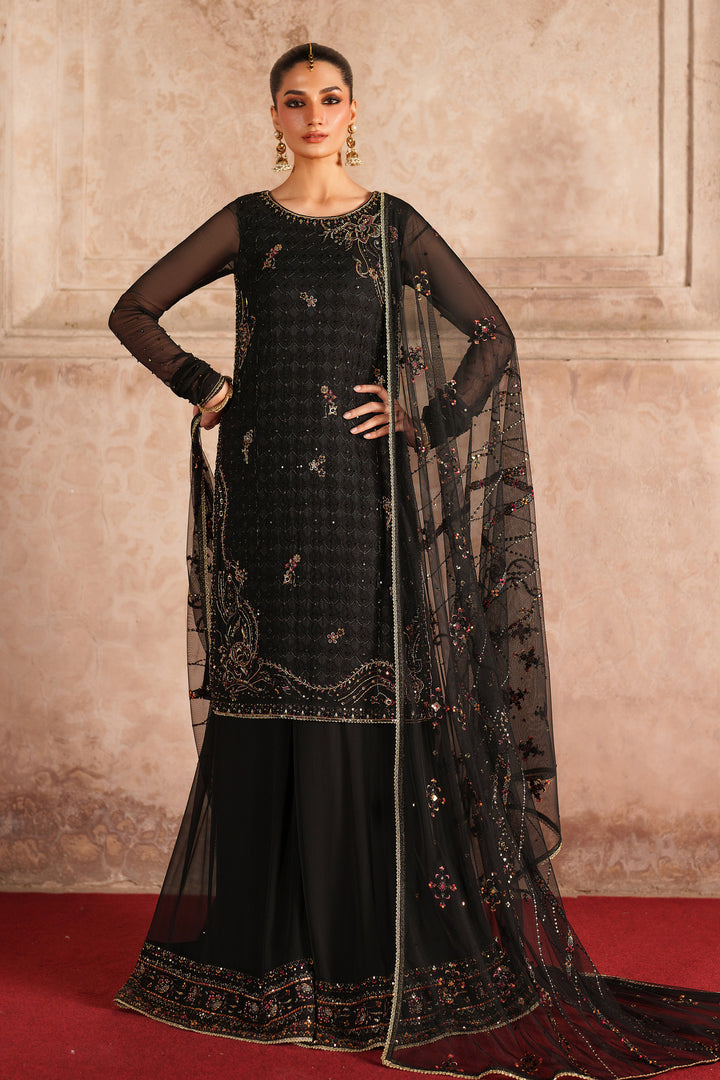Woman in a black embroidered traditional outfit with a sheer dupatta against a beige wall.
