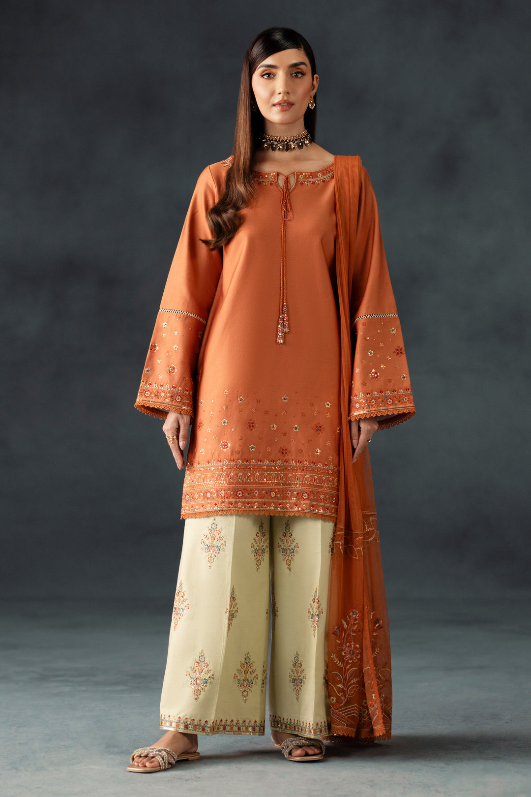 Woman wearing an orange embroidered outfit with beige pants against a dark background