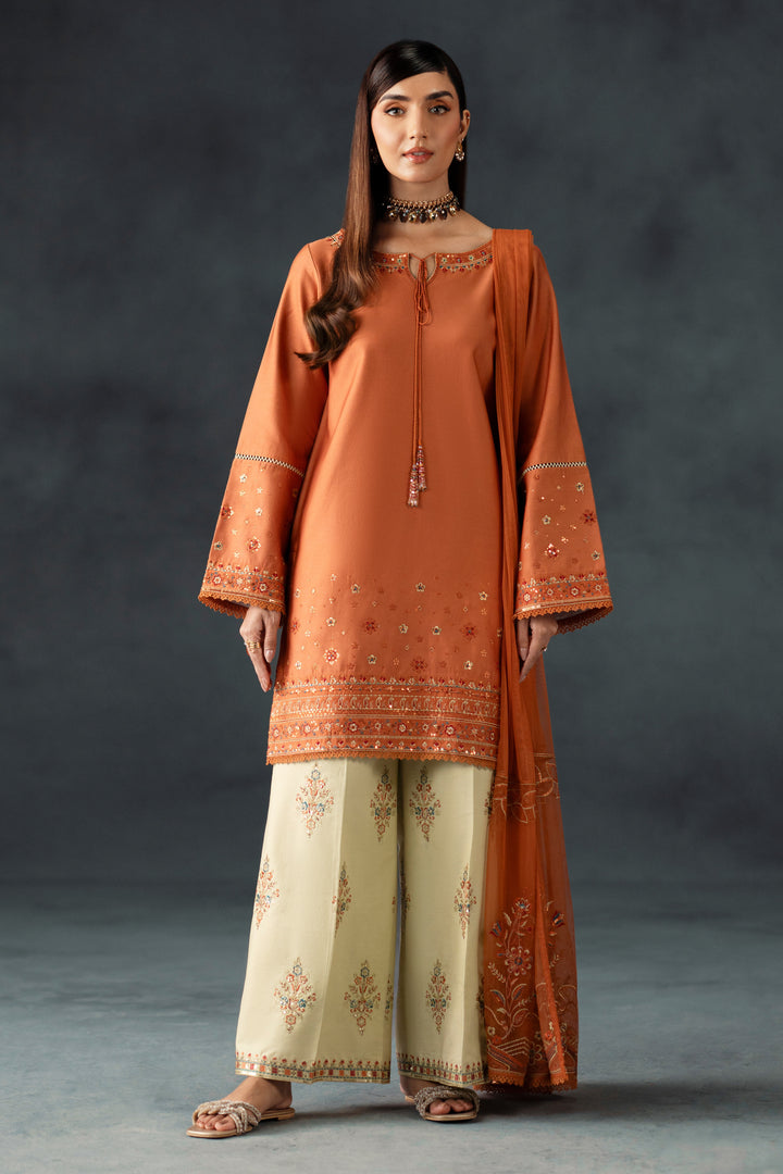 Woman wearing an orange embroidered outfit with beige pants against a dark background