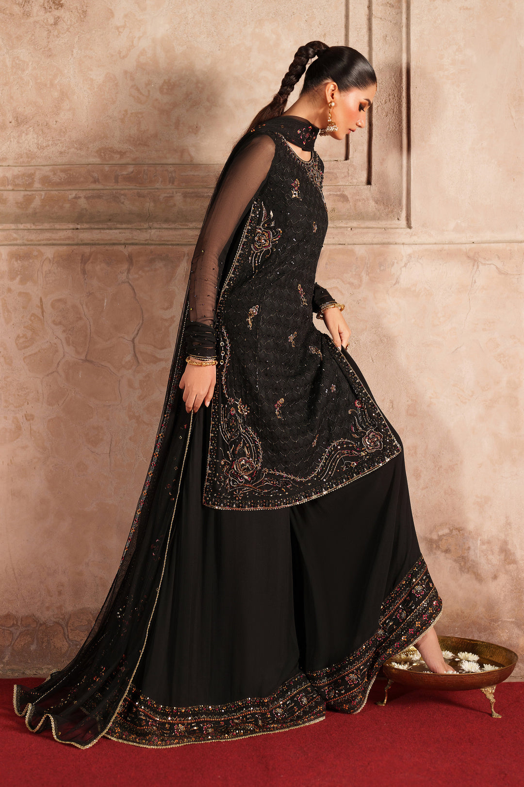 Woman in a black embroidered traditional outfit standing against a beige wall.