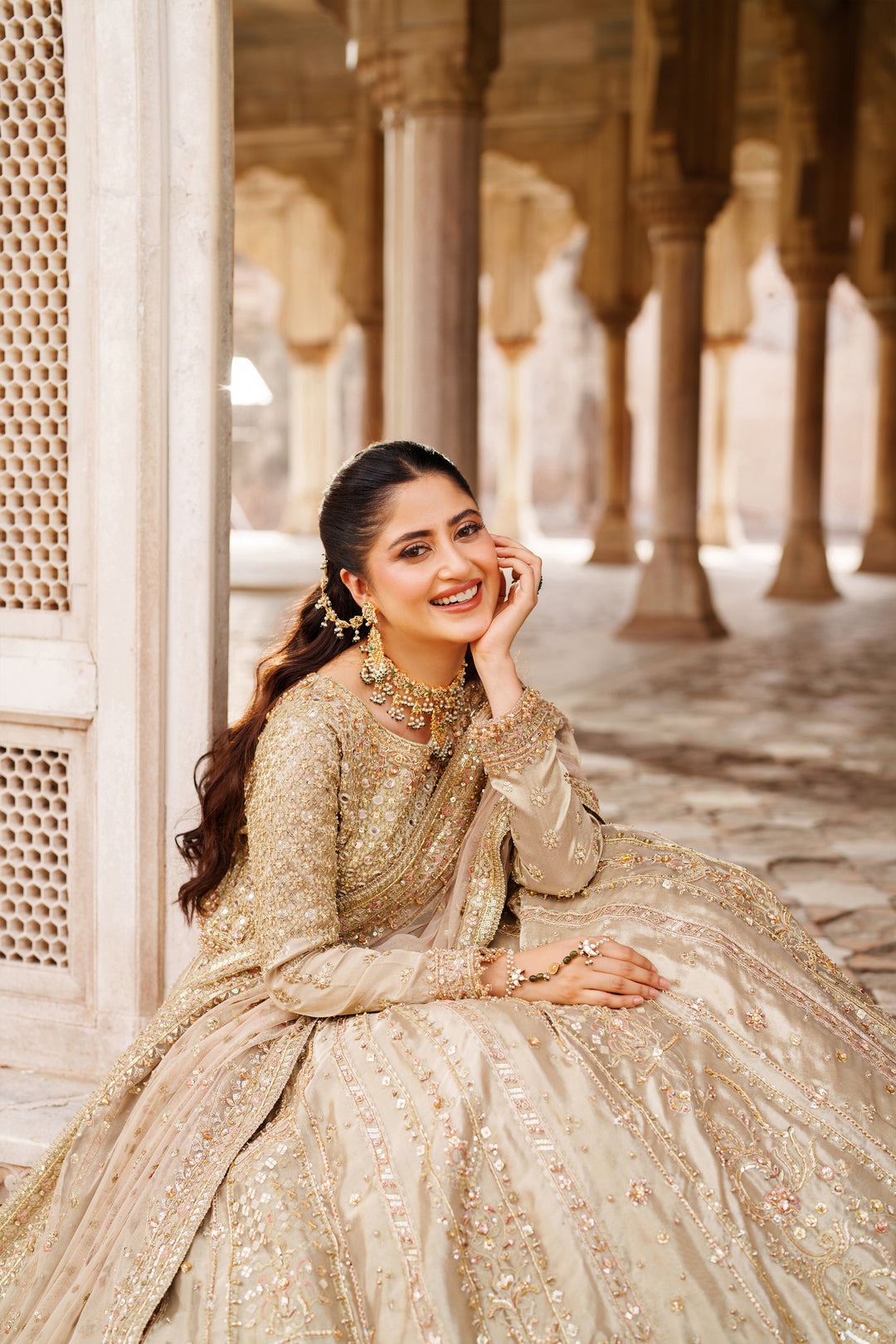 Styled sitting pose of NAQSHI Ruhab bridal gown in champagne gold with ornate embroidery and full skirt for wedding celebrations.