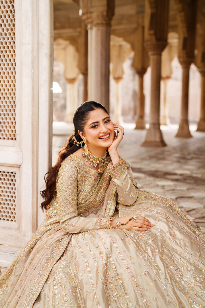 Styled sitting pose of NAQSHI Ruhab bridal gown in champagne gold with ornate embroidery and full skirt for wedding celebrations.