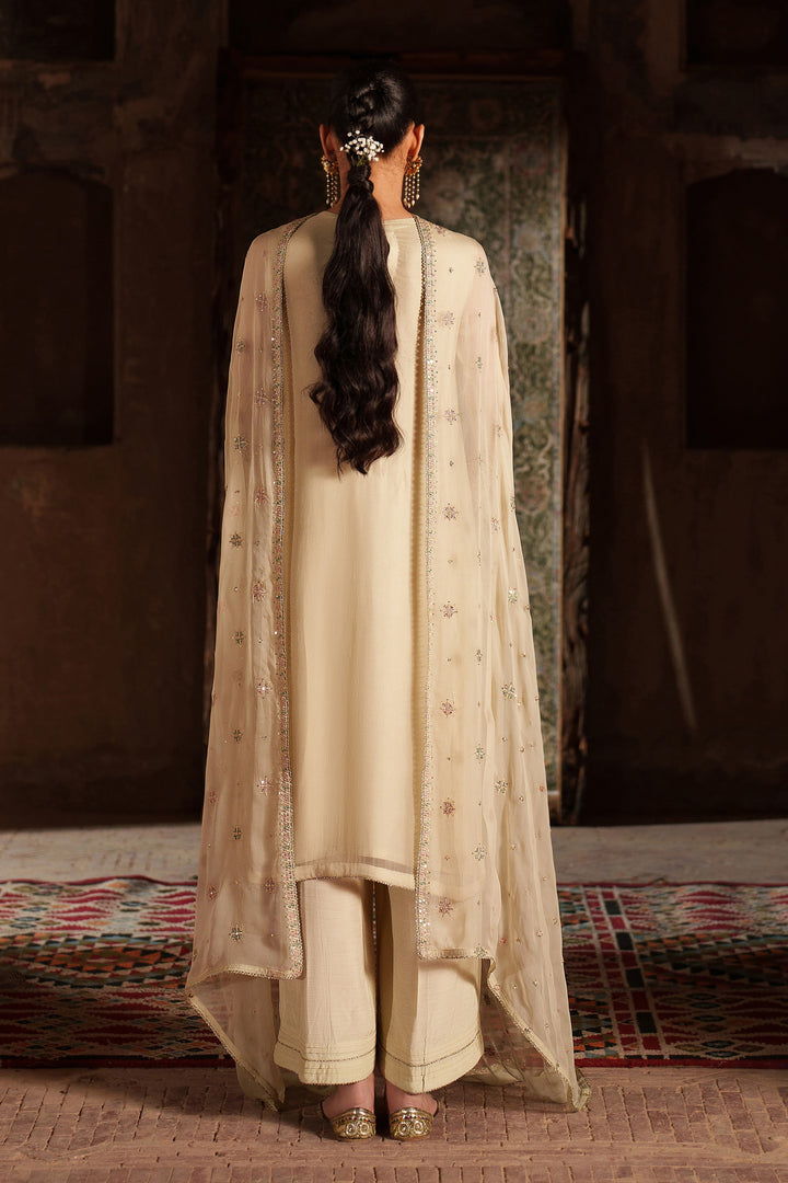 Woman in a cream traditional outfit with a long dupatta, standing in a warm-toned room.