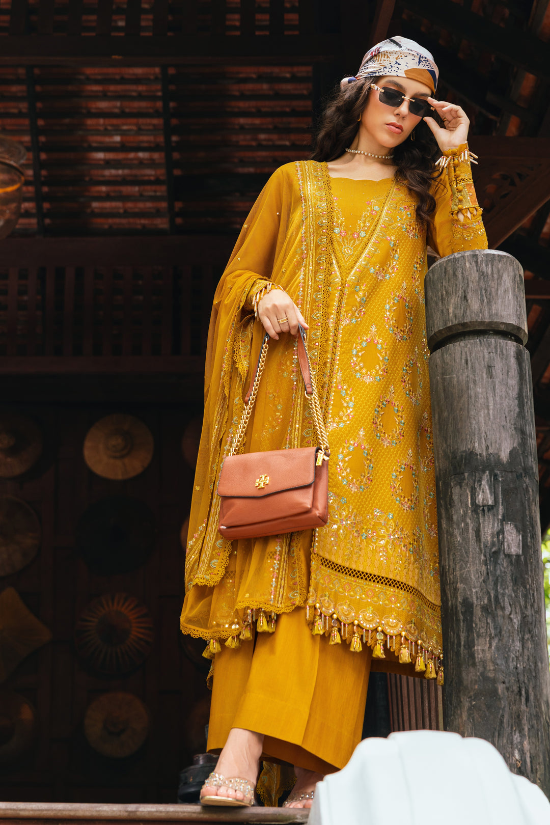 Woman in a yellow traditional outfit with sunglasses and a handbag, standing against a wooden background.
