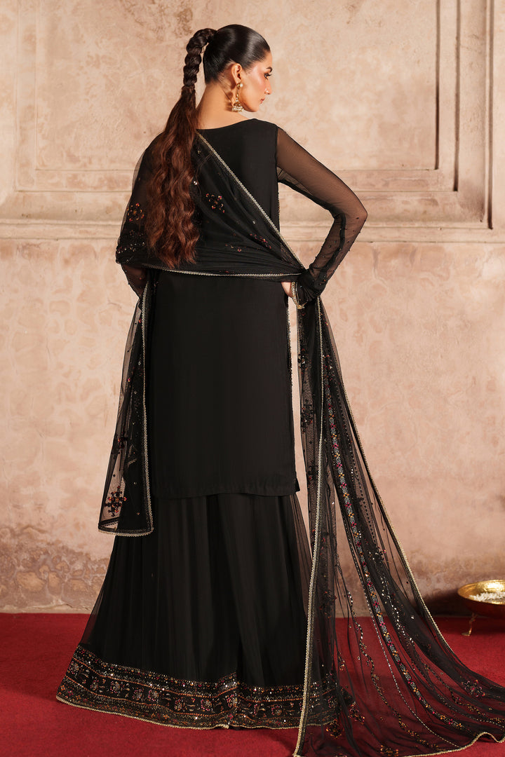 Woman in a black traditional outfit with a sheer dupatta against a beige wall.