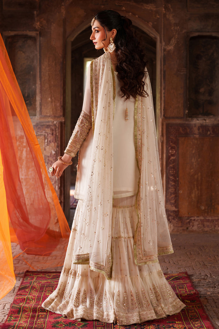 Woman in traditional white and gold outfit with orange dupatta in a decorative setting