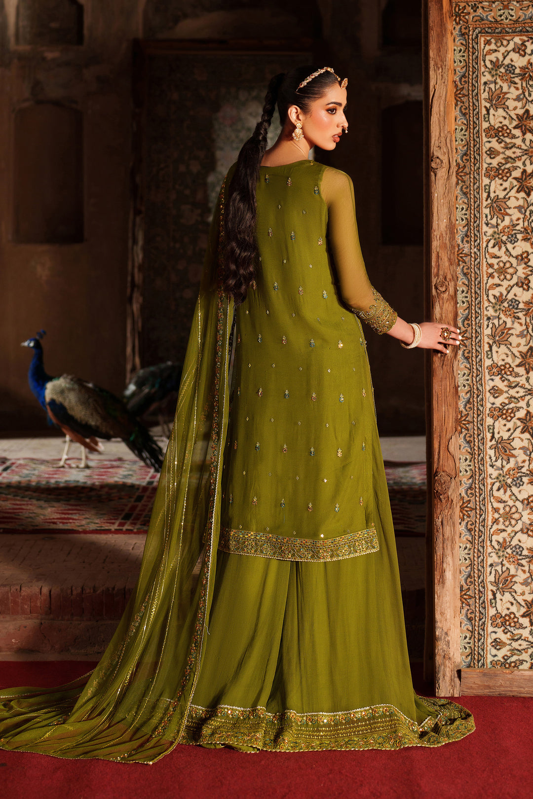Woman in a green traditional outfit standing in a doorway with peacocks in the background.