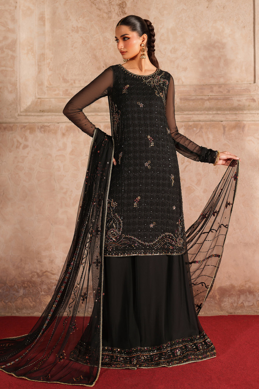 Woman wearing a black embroidered traditional outfit with a dupatta against a beige wall.