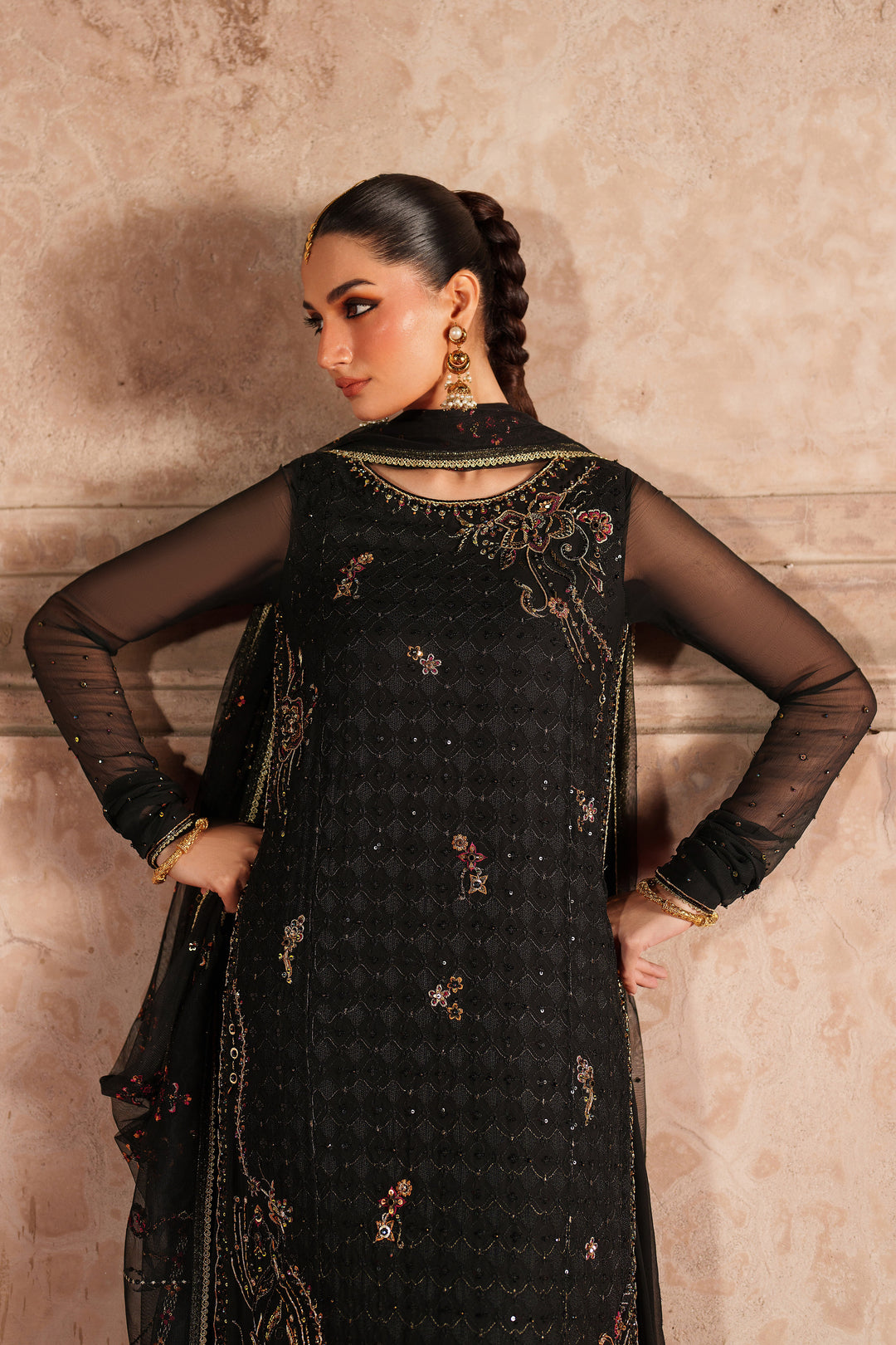 Woman wearing a black embroidered traditional outfit against a beige wall.