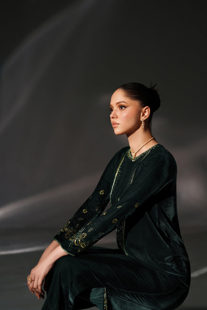 Woman in a dark green outfit with gold accents sitting against a dark background
