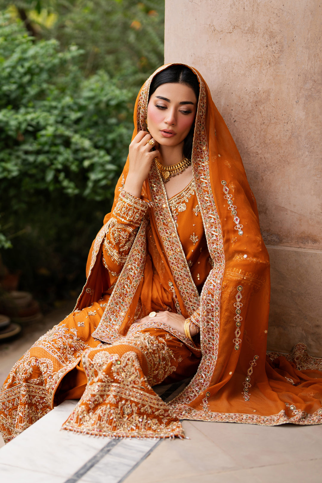 NAQSHI rust formal gown styled in seated pose, showing detailed hem border, panelled embroidery, full sleeves, and elegant fall of raw silk fabric.