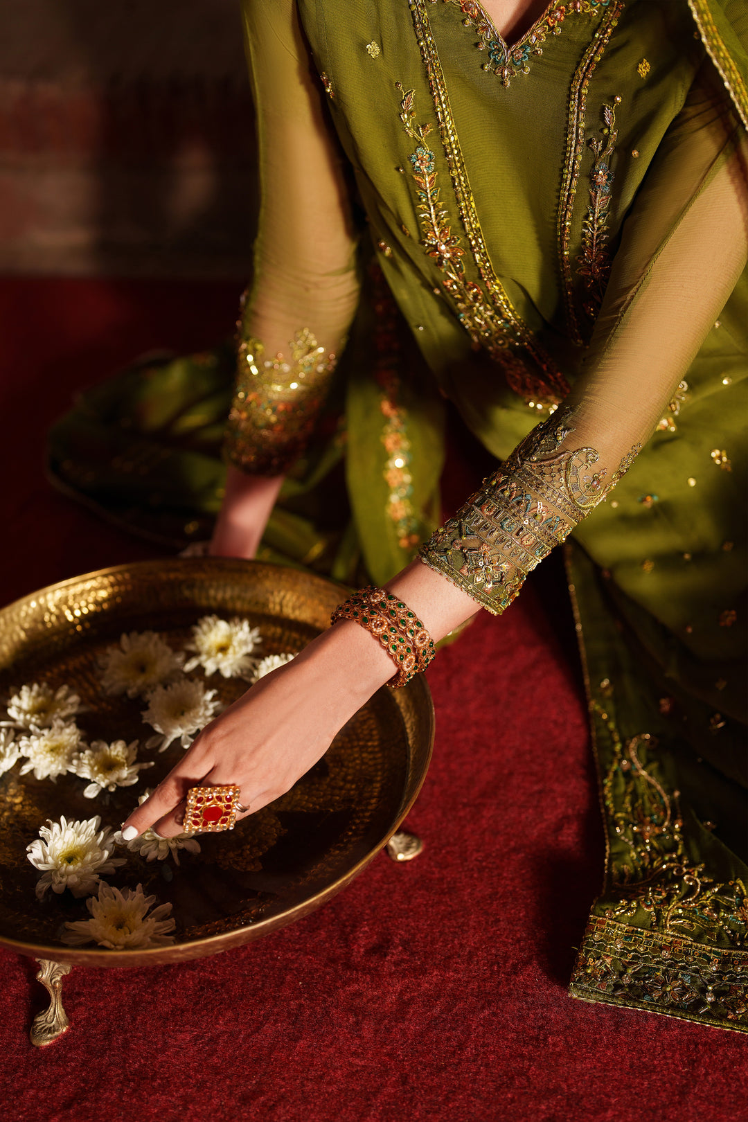 Woman in traditional green embroidered outfit with floral arrangement in a brass tray on a red surface