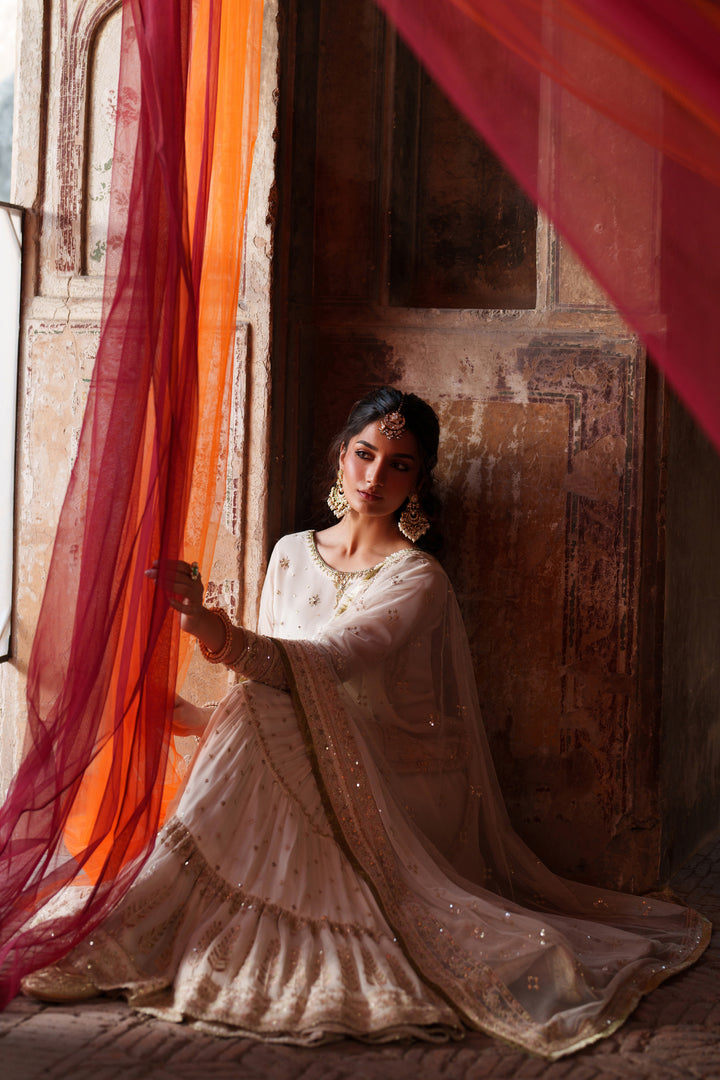Woman in a white traditional outfit sitting in a warm-toned room with red curtains.