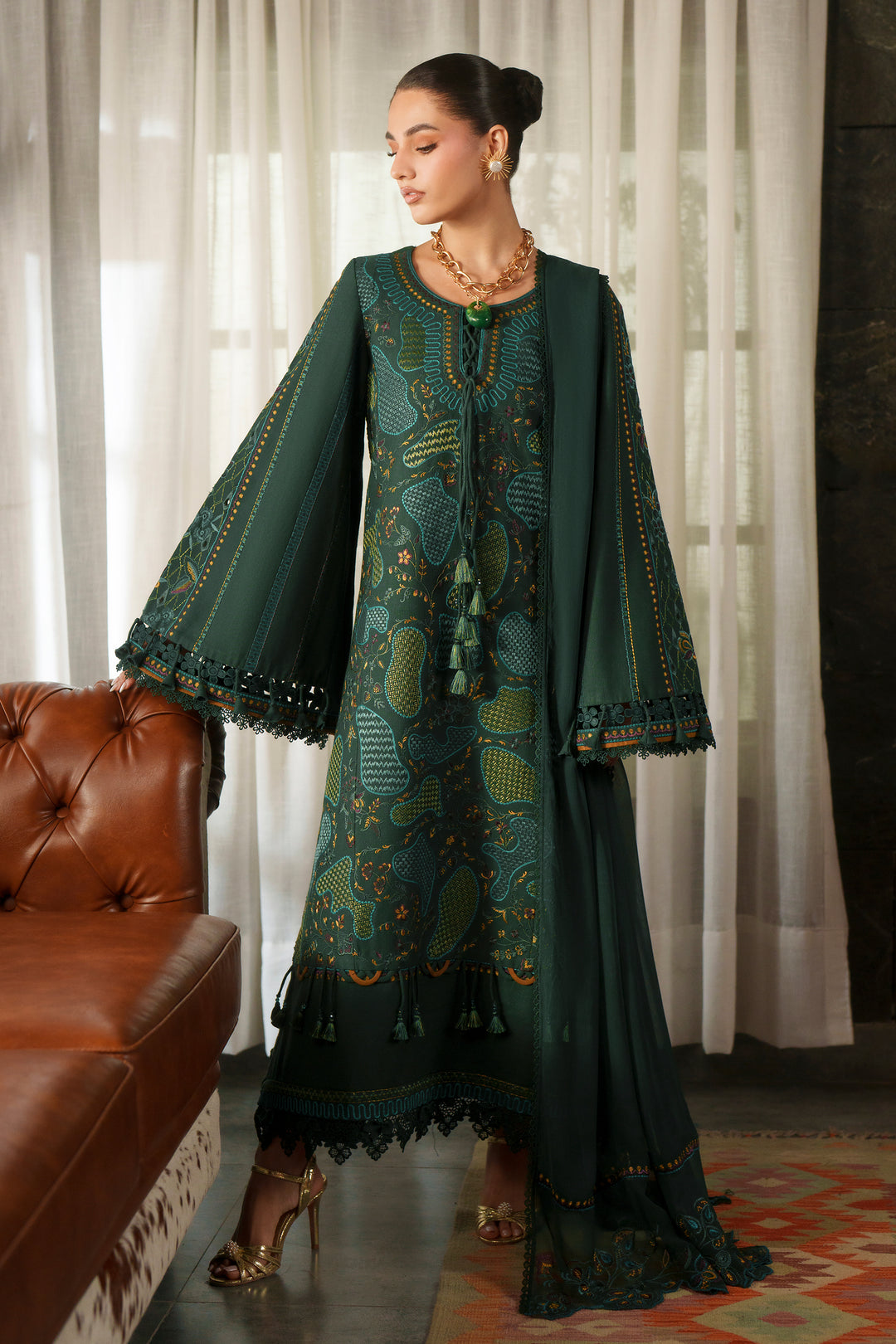 A woman models a dark green traditional outfit with intricate gold and teal embroidery, featuring a long kurta with wide bell sleeves and a matching dupatta, set in an indoor lifestyle environment with a brown leather sofa and white curtains.