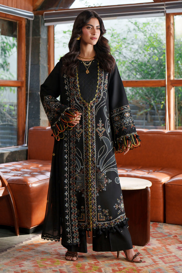 A woman models a long black embroidered kurta with gold and multicolor detailing, standing in a modern room with brown leather sofas and large windows.