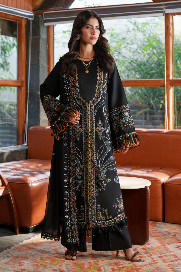 A woman models a long black embroidered kurta with gold and multicolor detailing, standing in a modern room with brown leather sofas and large windows.