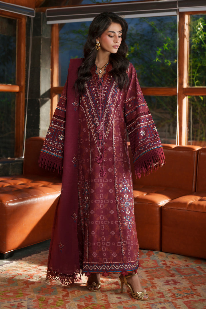 A woman models a long maroon embroidered traditional dress with wide sleeves and fringe details, standing in a furnished room with brown leather sofas and patterned rugs.