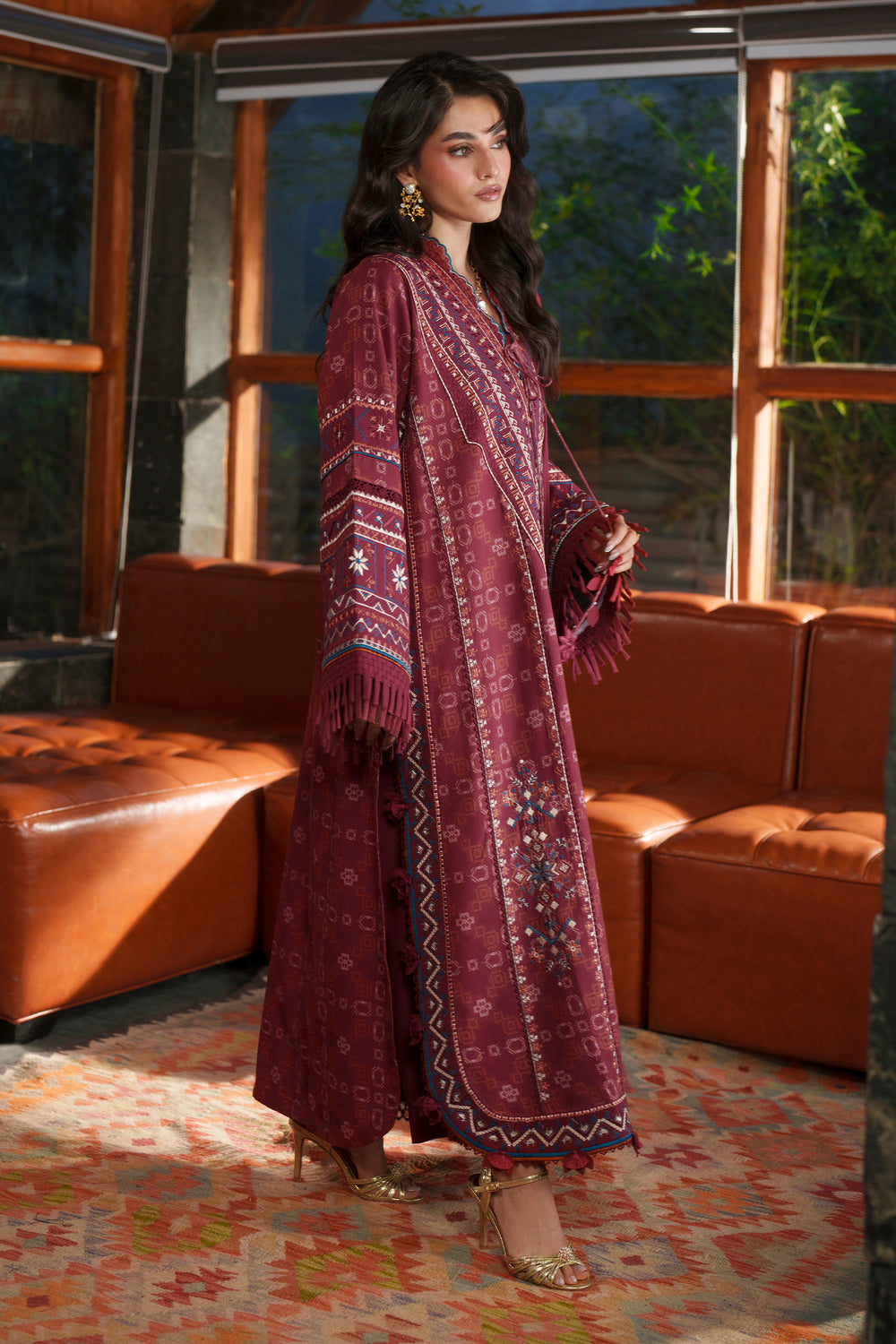 A woman models a traditional long-sleeve maroon ethnic outfit with intricate embroidery and fringe details, standing in a modern living room setting with leather sofas and large windows.