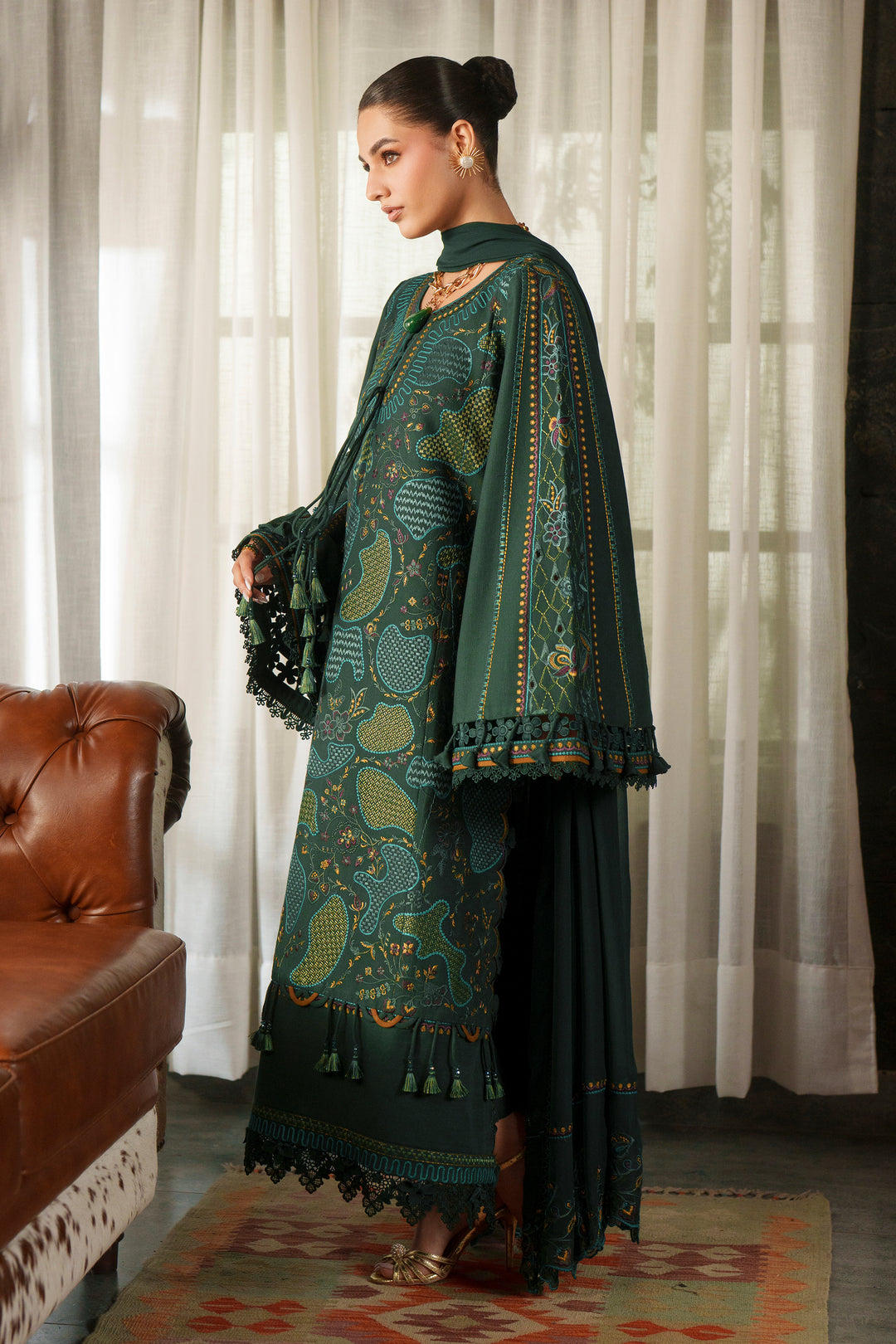 A woman models a dark green traditional outfit with intricate gold and teal paisley embroidery, a matching embroidered cape, and a long dark skirt, standing in a room with sheer white curtains and a brown leather sofa.