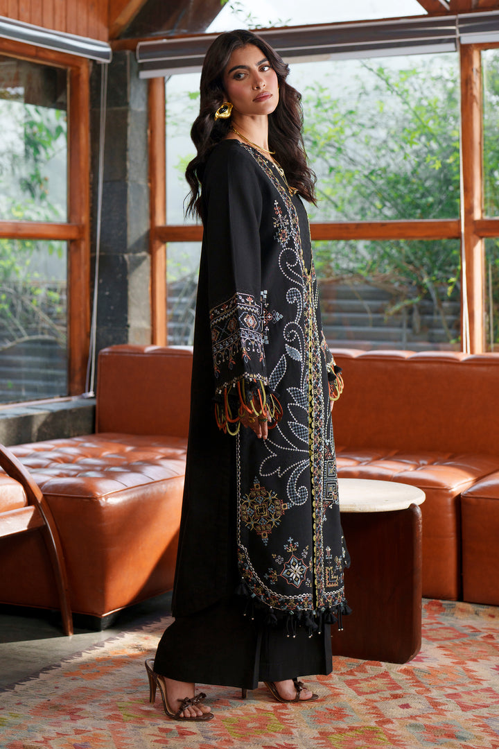 A woman models a black long kurta with intricate gold and silver embroidery, standing in a modern lounge room with brown leather sofas and large windows.