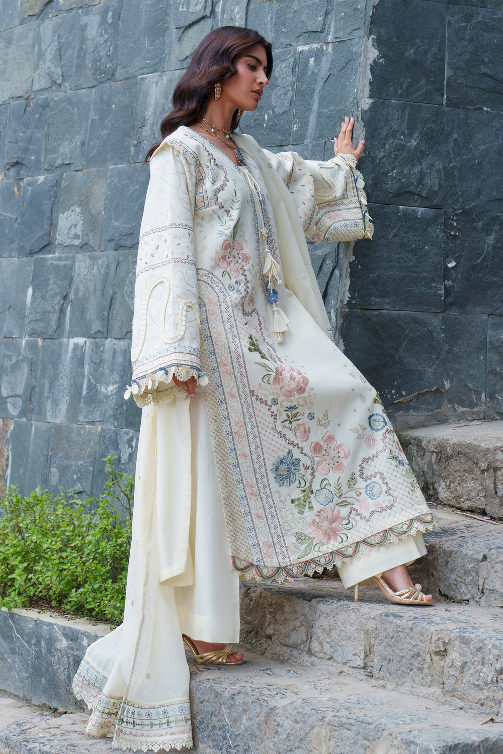 A woman models a traditional South Asian outfit consisting of a cream-colored embroidered kurta, matching trousers, and a dupatta, set against a dark gray stone wall and steps.