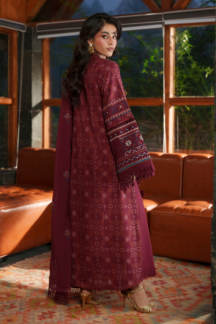 A woman wearing a long maroon embroidered kurta with matching dupatta and gold heels, standing in a modern room with brown leather sofas and large windows.