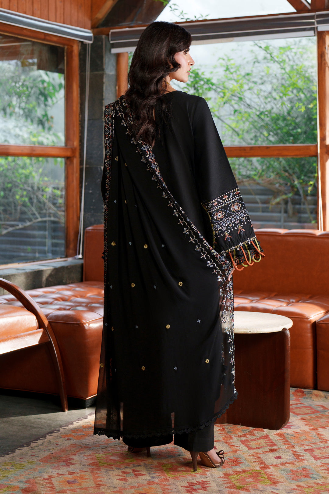 A woman stands with her back to the camera wearing a black traditional outfit with embroidered details and a matching dupatta in a furnished indoor space with large windows and brown leather seating.
