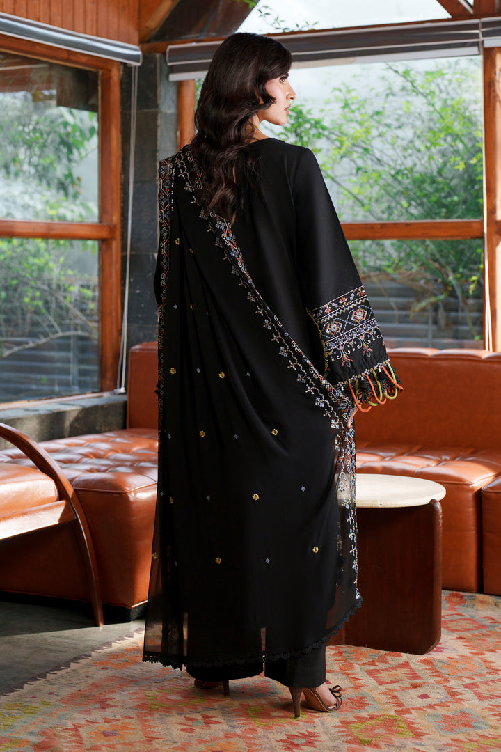 A woman stands with her back to the camera wearing a black traditional outfit with embroidered details and a matching dupatta in a furnished indoor space with large windows and brown leather seating.