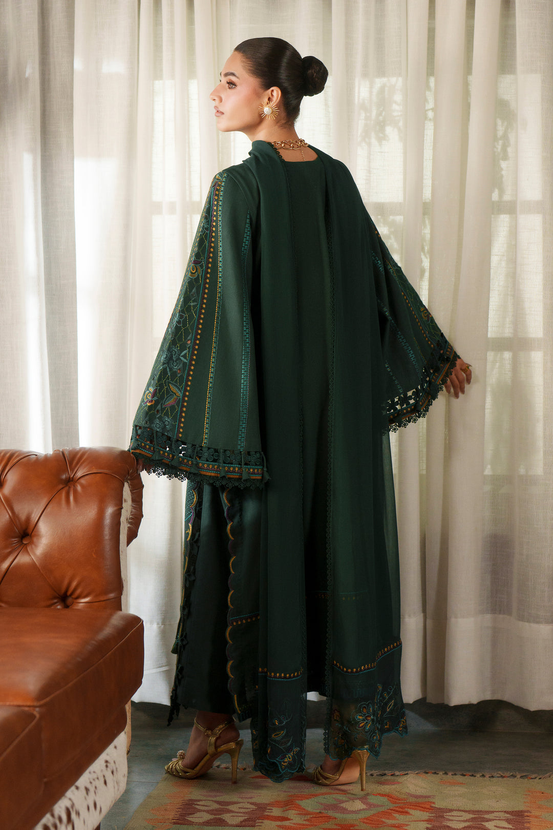 A woman wearing a dark green traditional outfit with wide embroidered sleeves and matching pants, standing indoors near sheer curtains and a brown leather sofa.
