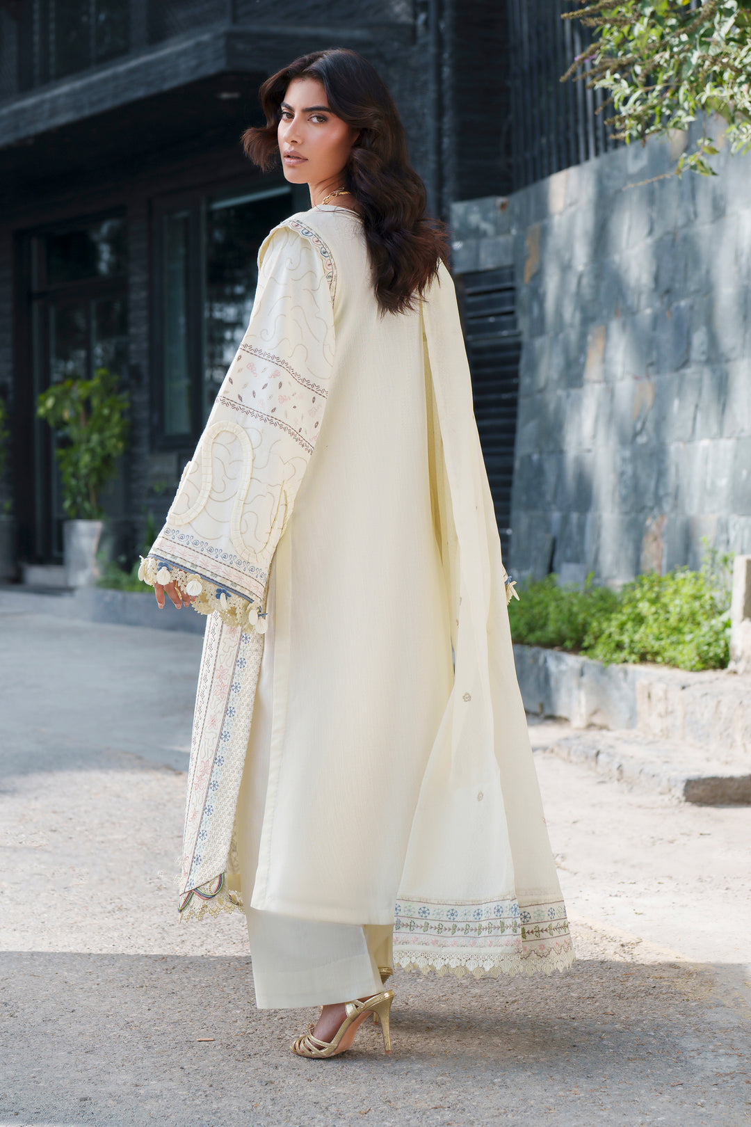 A woman wearing a cream-colored traditional outfit with embroidered details and a matching draped scarf, standing outdoors on a paved walkway.