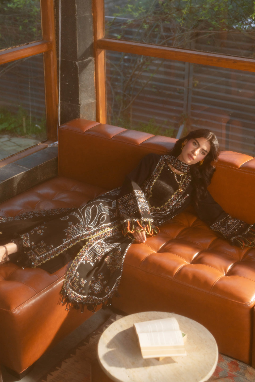 A woman reclines on a tufted orange-brown leather sofa in a sunlit interior, wearing a long black embroidered dress or robe with ornate gold and white detailing. A small round table with an open book sits in front of the sofa, and large windows with wooden frames are visible behind her.