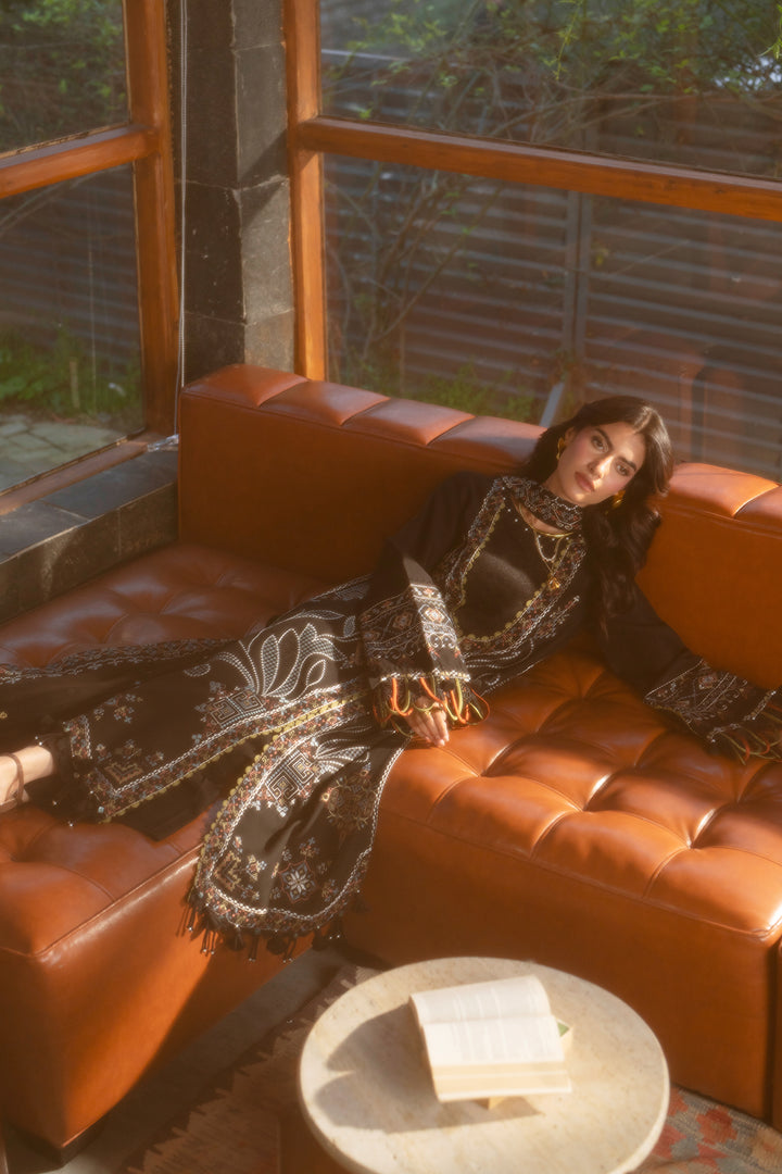 A woman reclines on a tufted orange-brown leather sofa in a sunlit interior, wearing a long black embroidered dress or robe with ornate gold and white detailing. A small round table with an open book sits in front of the sofa, and large windows with wooden frames are visible behind her.