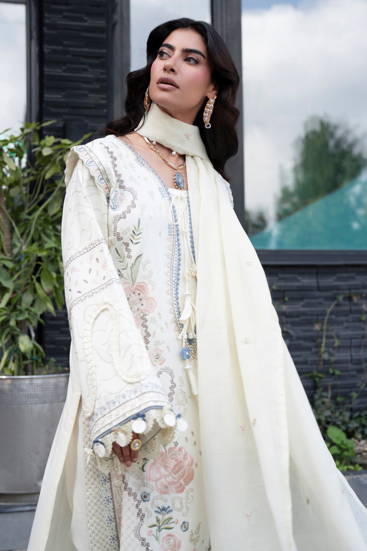 A woman wearing a cream-colored embroidered traditional outfit with floral patterns and a matching draped scarf, photographed outdoors in front of a modern building with plants.