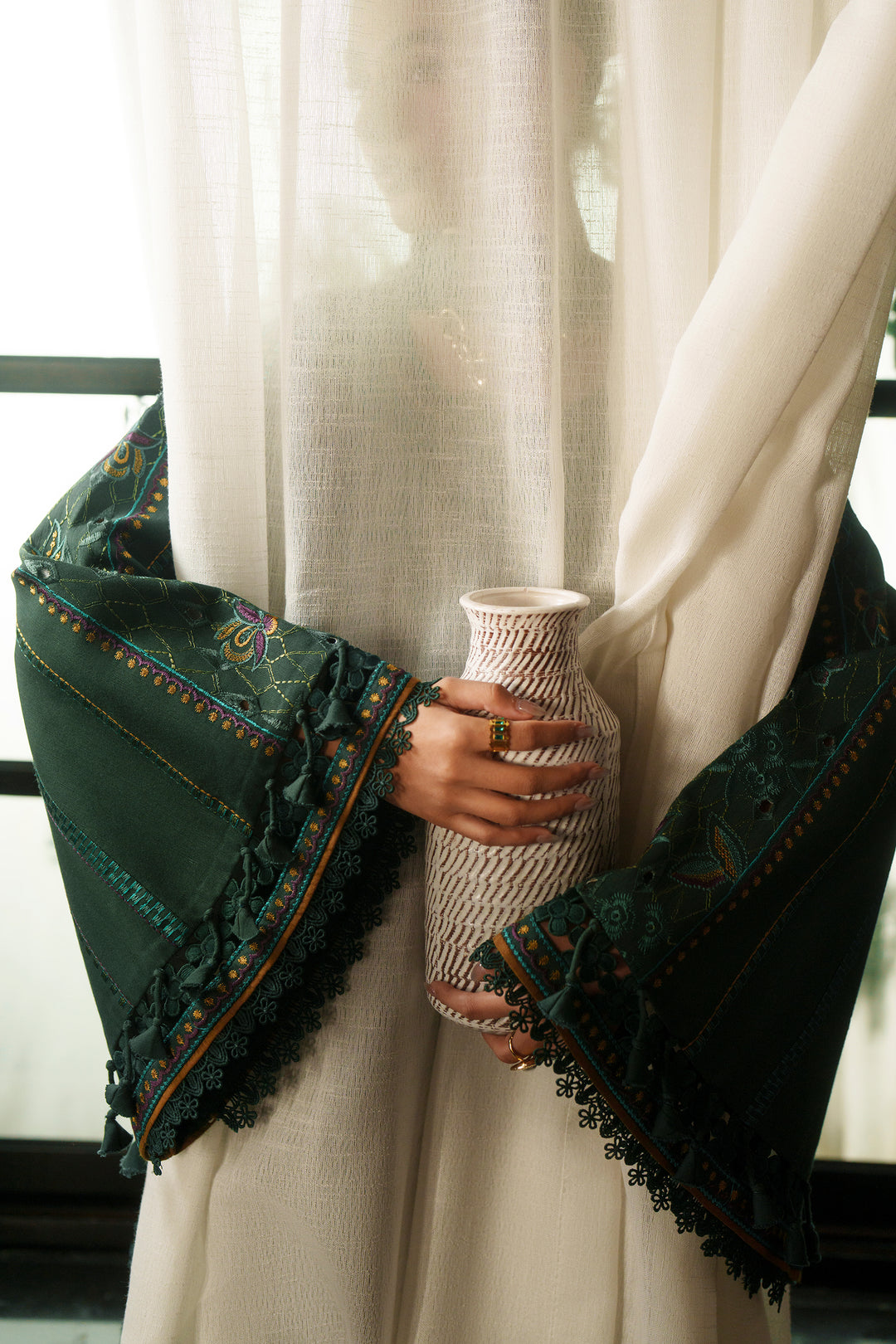 A person wearing a cream-colored traditional outfit with dark green embroidered sleeves holds a small woven vase in front of sheer white curtains.