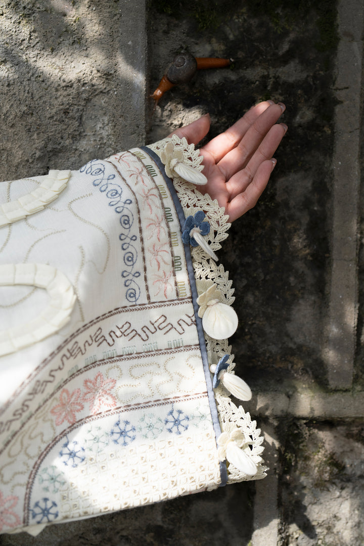 A close-up of a hand resting against a weathered stone wall, wearing a cream-colored sleeve decorated with blue and pink floral embroidery, lace trim, and white shell-like buttons.
