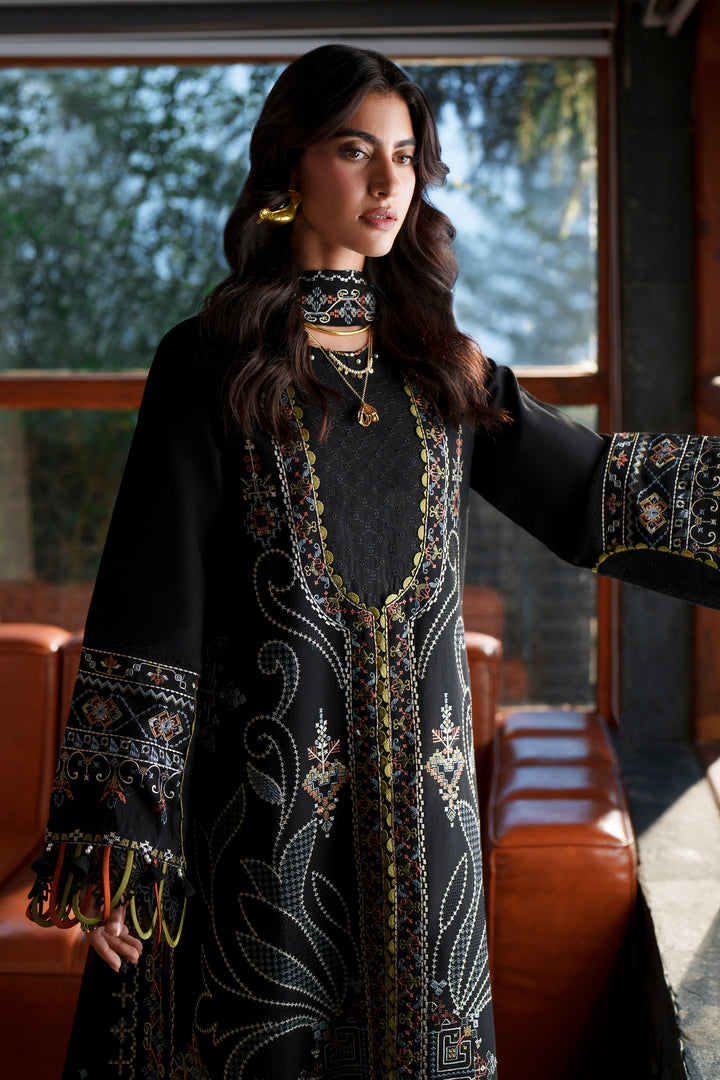 A woman wearing a black embroidered traditional outfit with intricate gold, white, and red patterns, standing in a modern interior with large windows and orange-brown seating.